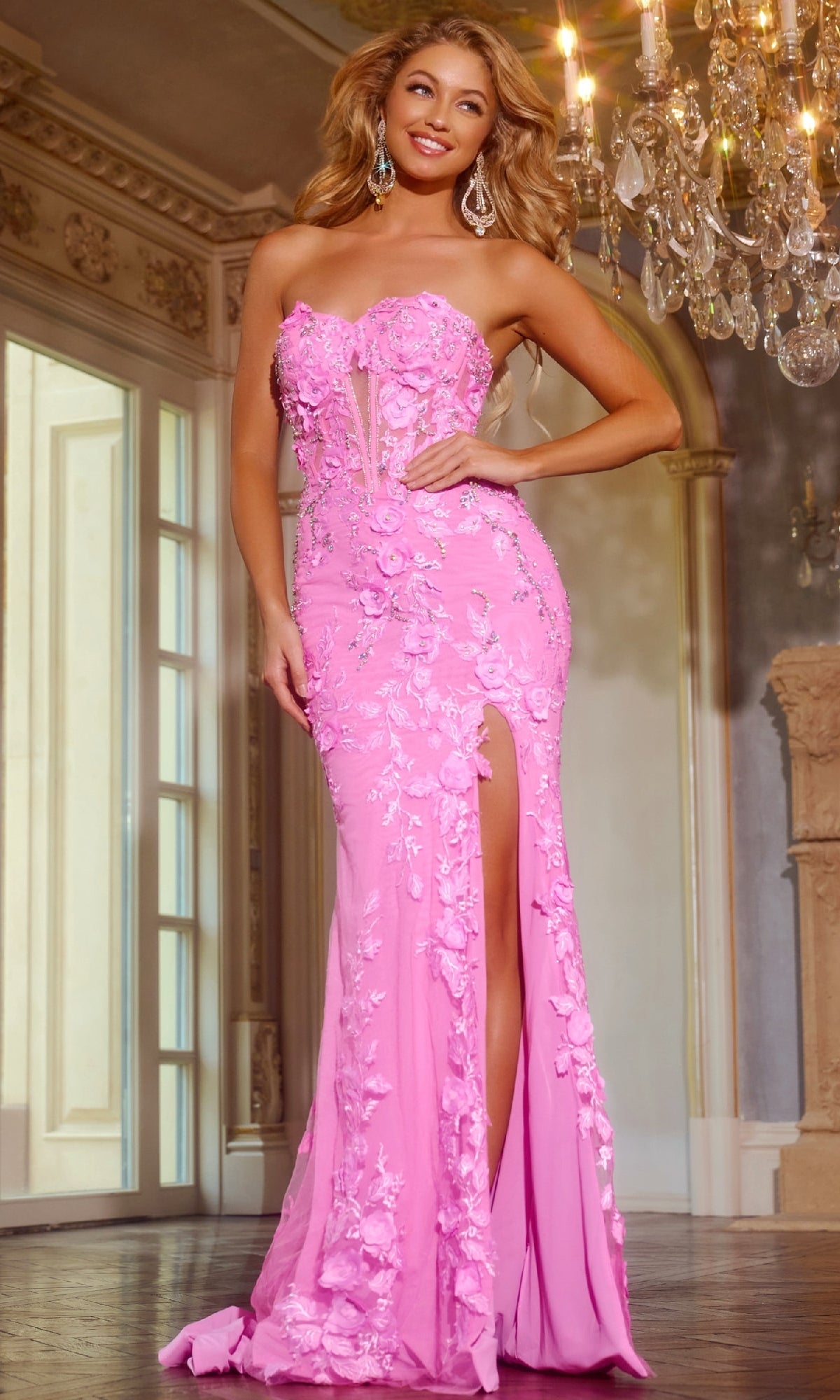 A woman stands indoors near a chandelier, wearing the Jovani D5707 long pageant dress—strapless, pink, with floral embellishments and a thigh-high slit.